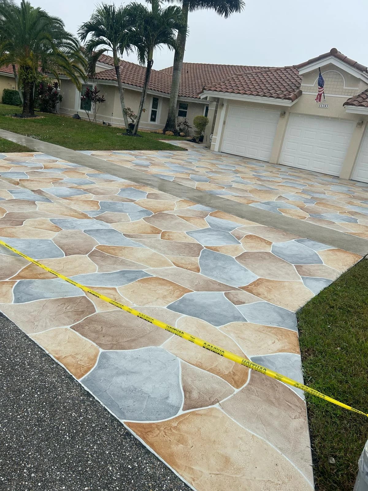 Ground-level view of a completed multi-tone tan, gray, and blue flagstone stamped concrete driveway with caution tape marking the fresh curing surface